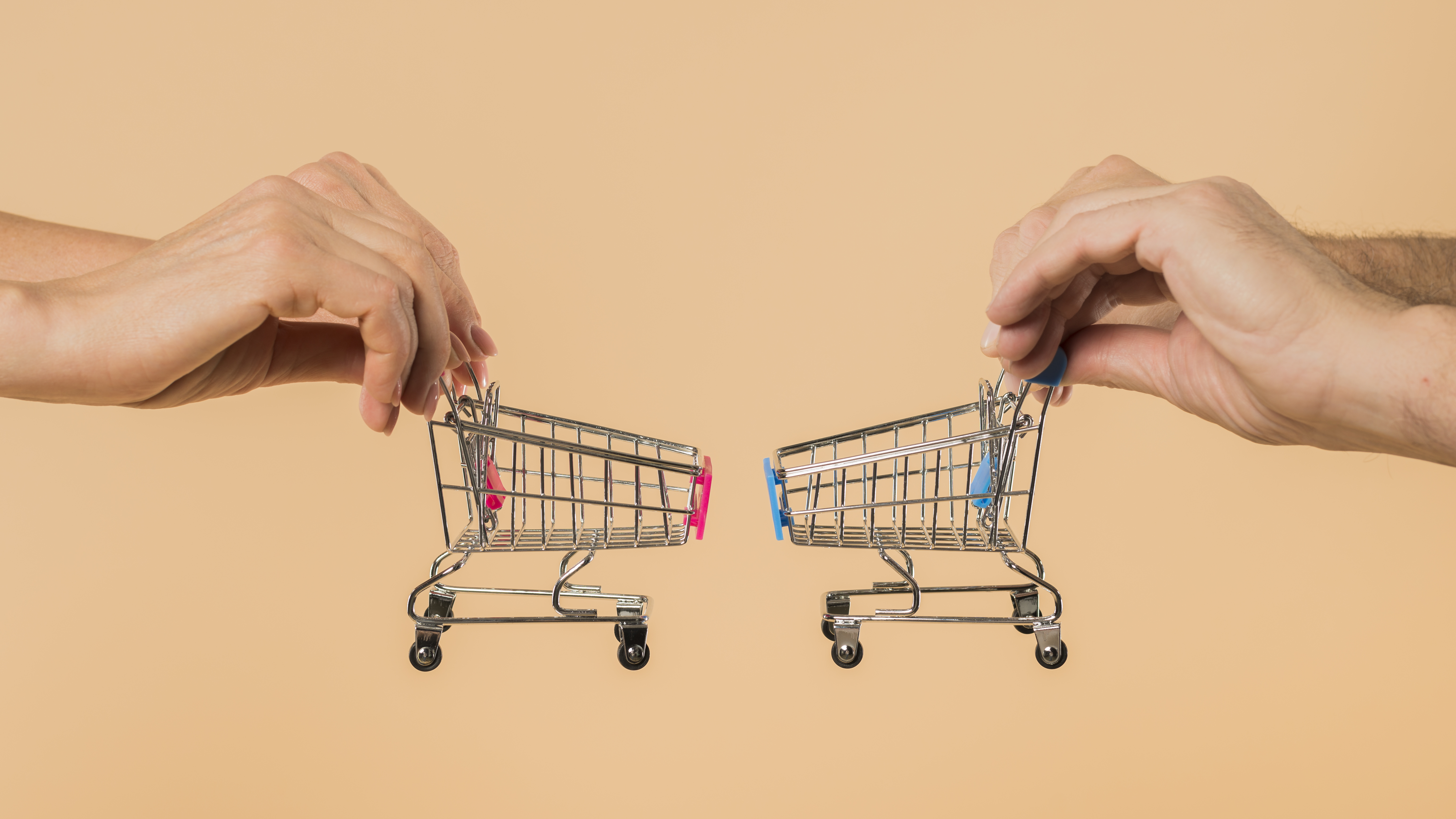 Pares de mãos seguram mini carrinhos de supermercado indo ao encontro um do outro. Fundo alaranjado