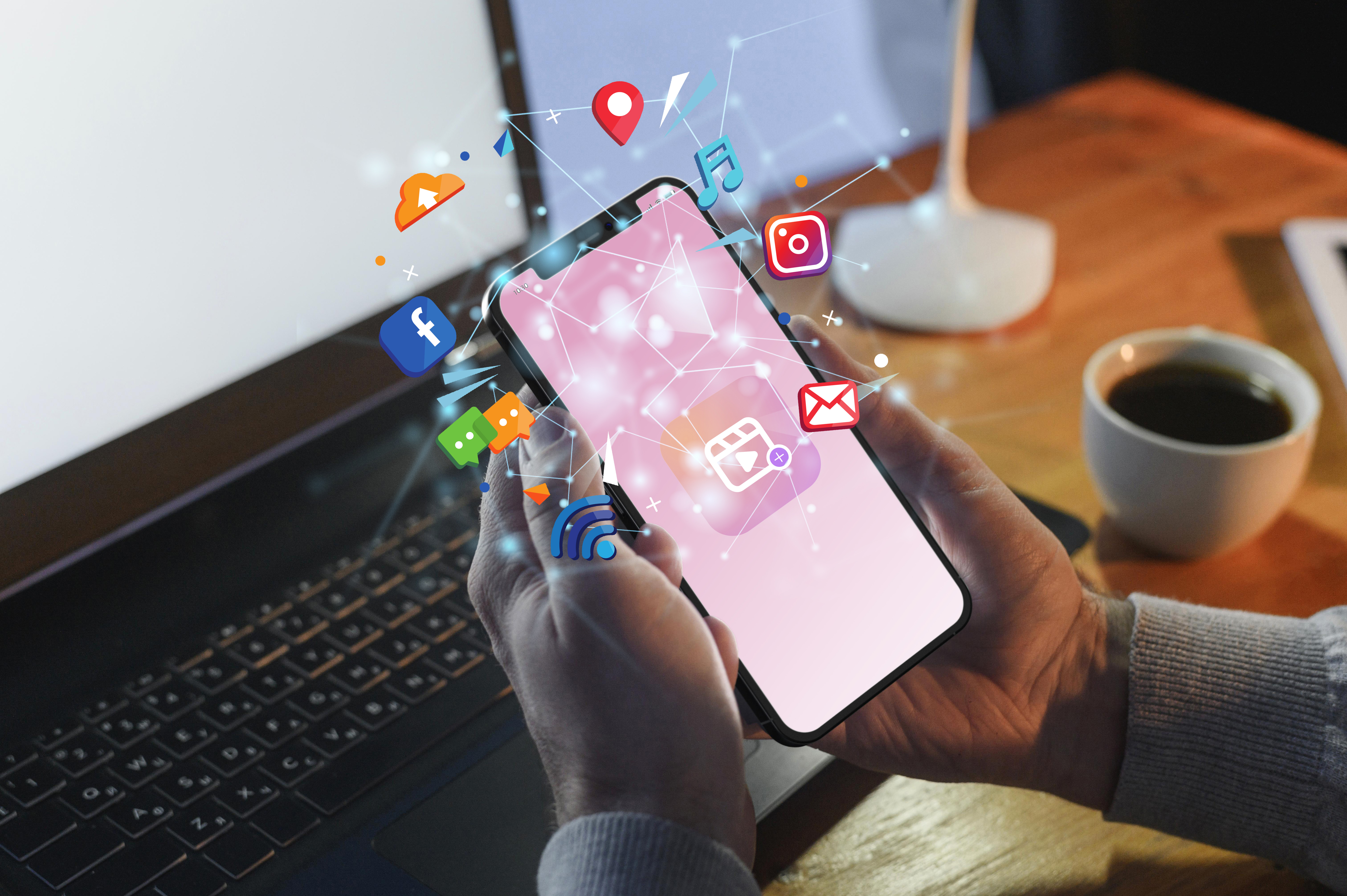Na imagem, há uma mão masculina segurando um celular com vários ícones de canais digitais diferentes saindo dele em forma de ilustração. Em frente a mão há um notebook e ao lado uma xícara de café e luminária