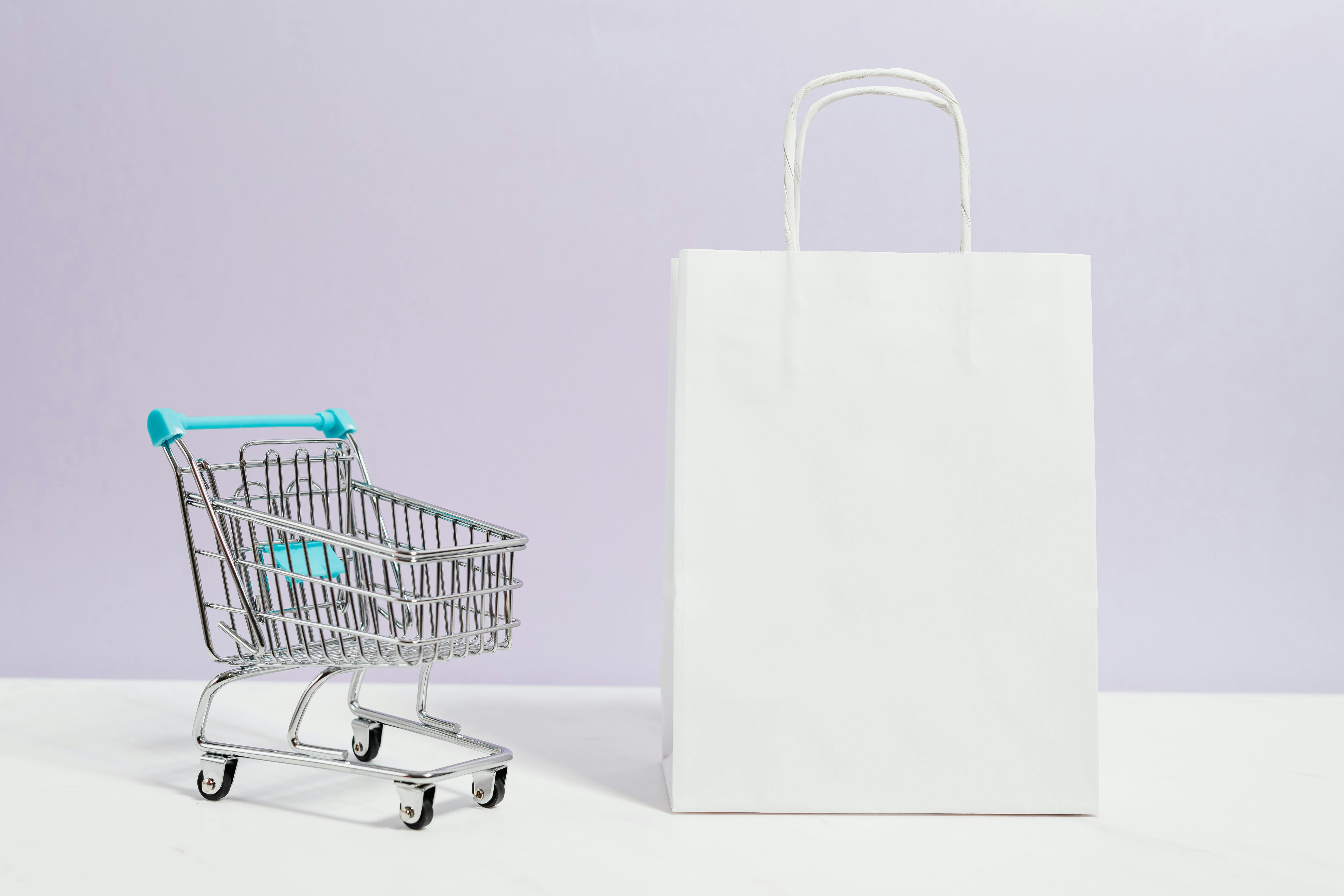 Na foto, há um mini carrinho de supermercado ao lado de uma sacola de compras e fundo lilás