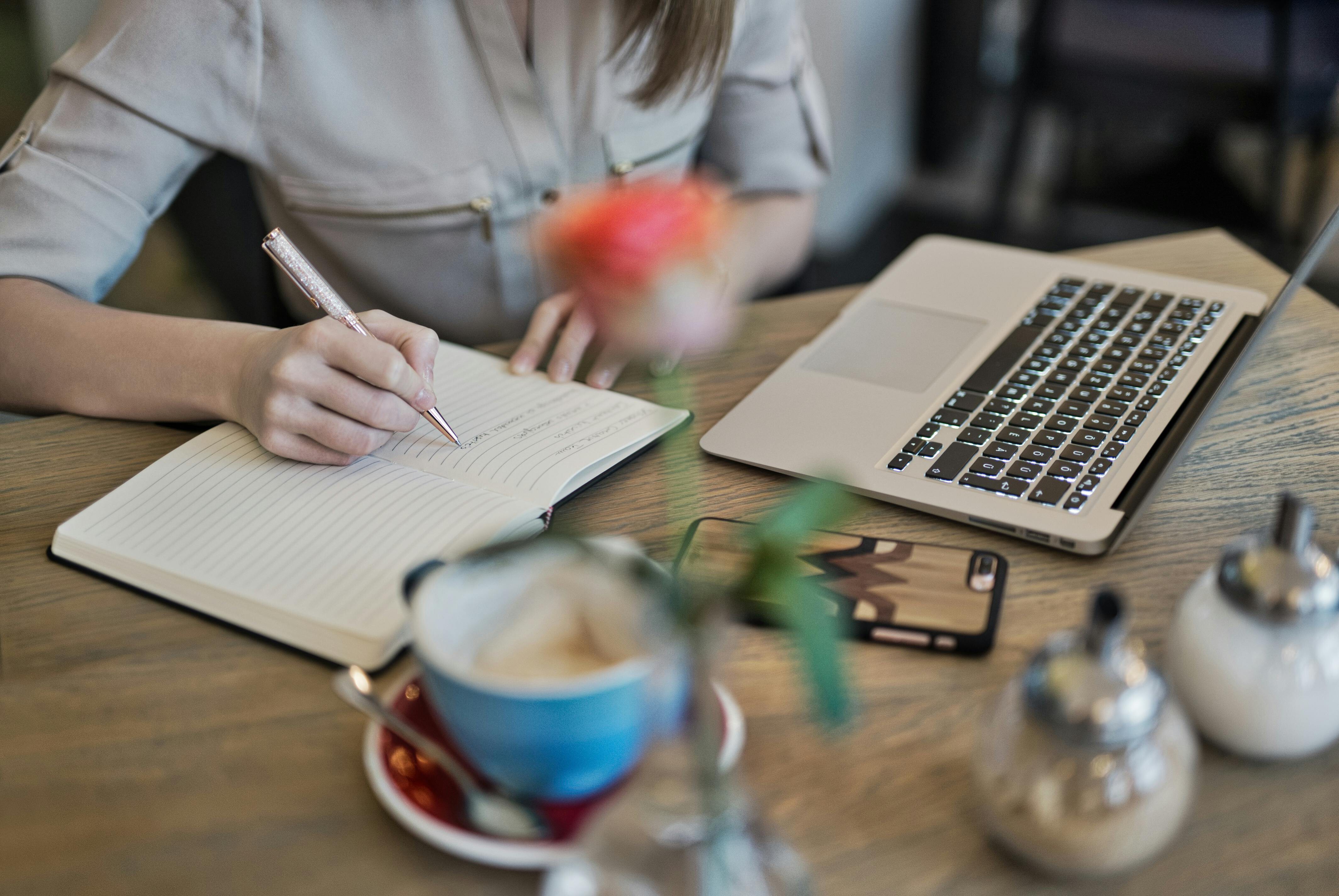 Imagem de uma mulher em frente a um notebook fazendo anotações em um caderninho. Ao lado tem um café e em sua frente, um celular